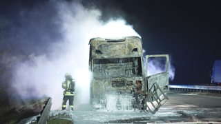 Ein Feuerwehrmann steht neben einem ausgebrannten und immer noch rauchenden Lkw.