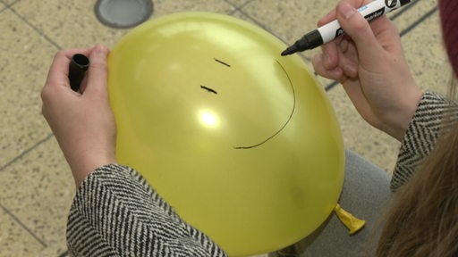 Eine Person malt ein Gesicht auf einen gelben Luftballon.