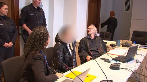 Bei einem Gerichtsprozess unterhält sich der Verteidiger mit einem Polizisten, während sich die Angeklagte auf einen Stuhl setzt.