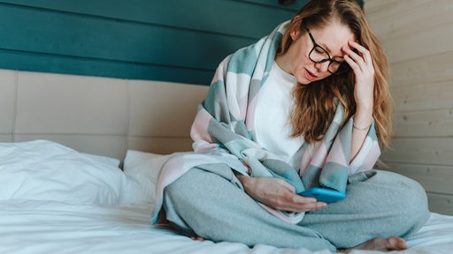 Eine junge Frau sitzt auf dem Bett, schaut aufs Handy und hält sich dabei den Kopf.