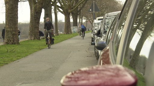 Fahrradfahrer auf der linken Seite, rechts parkende Autos. 