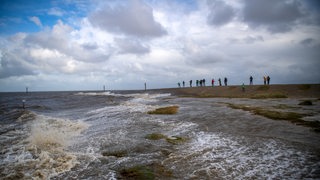 Wellen schlagen an den Nordseestrand, im Hintergrund stehen Menschen auf einem Damm.