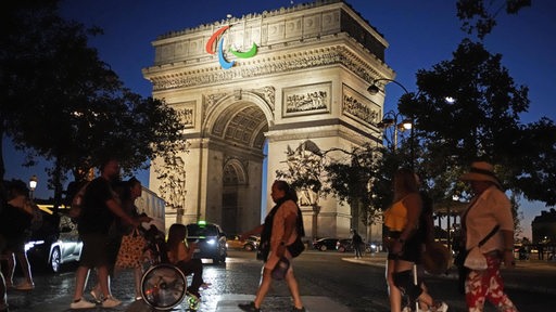 Blick am Abend auf den angestrahlten Arc de Triomphe in Paris, an den das Logo der paralympischen Spiele angebracht ist.