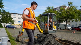 Ein Bauarbeiter in Warnweste steht vor einem Bagger auf einem Parkplatz.