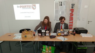 Zwei Männer hinter zusammengerückten Tischen vor dem Logo der Partei "Die Partei"