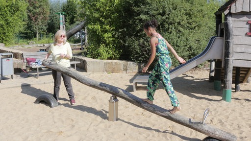 Ein Kind balanciert über eine Wippe, eine Frau hält die Wippe fest.