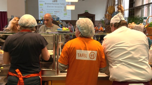 Drei Personen stehen hinter einem Tresen, man sieht ihre Rücken. EIne trägt ein Shirt mit der Aufschrift "Tafel".