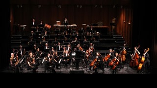 Musiker sitzen als Orchester mit ihren Instrumenten auf einer erleuchteten Bühne.