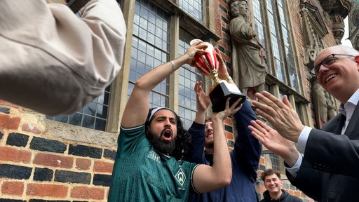 Zwei Männer halten einen Pokal in die Höhe. Daneben steht Andreas Bovenschulte. 