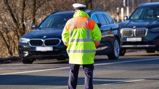 Ein Mann in Polizeiuniform steht mit dem Rücken zur Kamera auf einer Straße. Im Hintergrund sind zwei schwarze BMWs zu sehen.