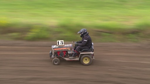 Ein junger Mann fährt auf einem Rasenmähertraktor über eine schlammige Rennbahn