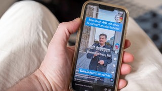 Hand hält ein Mobiltelefon auf dessen Display auf der Plattform TikTok ein Video des AFD Politikers Max Krah zu sehen ist, der Schüler dazu auffordert sich zur AFD zu bekennen.