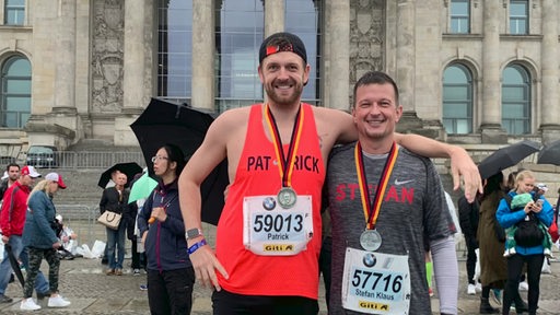 Zwei Männer stehen mit Laufkleidung, Startnummern und Medaillen vor dem Reichstag