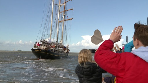 An einem Pier stehende Menschen winken einem abfahrenden Segelschiff.