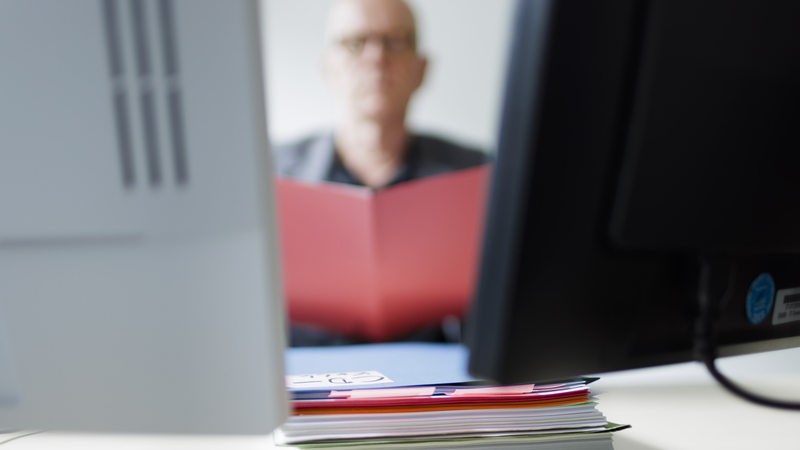 Ein Mann sitzt hinter Computerbildschirmen und liest in einem Ordner.
