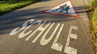 Ein dreieckiges Schild mit Kindern und Schriftzug "Schule" auf einer Straße vor einer Dorfschule.