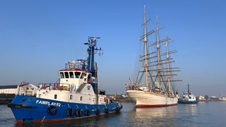 Ein Segelschiff wird von zwei Schlepper geschleppt.