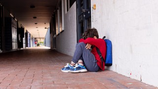 Ein Grundschüler sitze mit seinem Rucksack, den Kopf zwischen den Armen, vor der Schule.