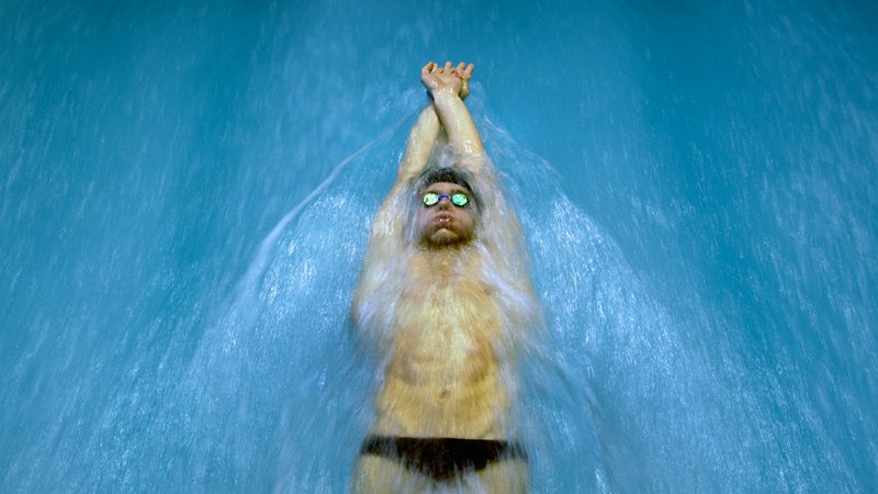 Ein Mann mit Badekappe und Schwimmbrille schwimmt auf dem Rücken durch ein Wasserbecken.