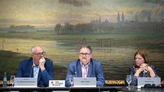 Das Bild zeigt drei Menschen, Bürgermeister Andreas Bovenschulte, Finanzsenator Björn Fecker sowie Wirtschaftsministerin Kristina Vogt, die gemeinsam an einem Tisch mit Mikrofonen vor einem landschaftlichen Gemälde, das Bremen zeigt.
