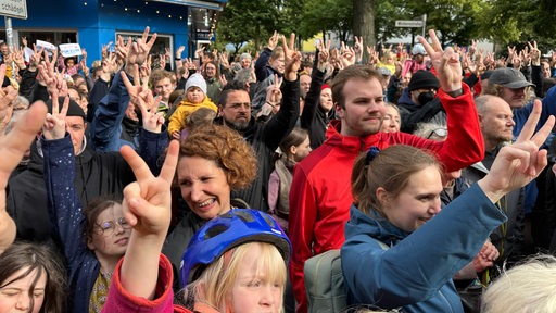 Zahlreiche Menschen haben sich auf einer Straße versammelt und heben eine Hand