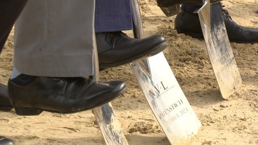 Drei Personen stehen während der Einweihung einer Baustelle auf Spaten