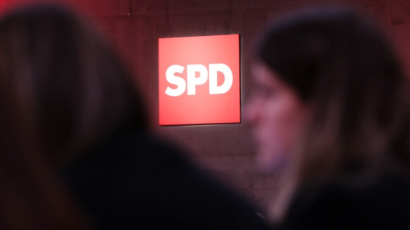 Das Logo der SPD leuchtet im Hintergrund, im Vordergrund sind Menschen unscharf zu sehen.