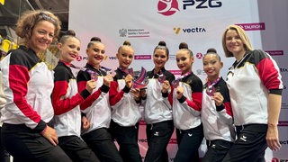 Sechs jungen Sportlerinnen tragen Traininganzüge und präsentieren ihre Silvermedaillen. Umrahmt werden sie von ihren beiden Trainierinnen-