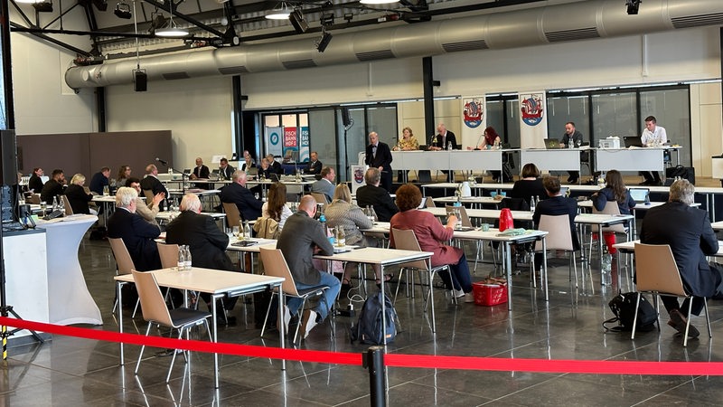 Zahlreiche Menschen in einem Saal sitzen an Tischen.