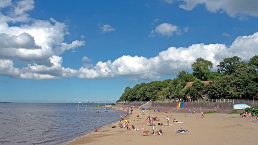 Links das Meer, rechts ein Sandstrand, mehrere Menschen liegen dort auf Strandhandtüchern.