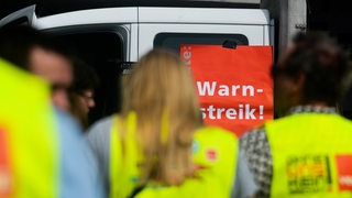 Menschen in gelben Warnwesten stehen vor einem roten Schild mit der Aufschrift "Warnstreik!"