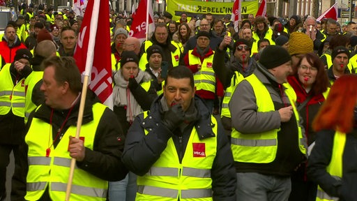 Mehrere Menschen sind in gelben Warnwesten zu sehen. Manche halten eine Fahne nach oben wo "verdi" draufsteht.