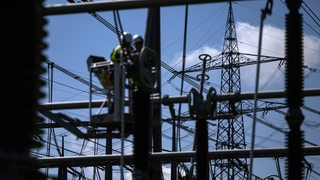 Bauarbeiter arbeiten an einem Umspannwerk. Im Hintergrund ist ein Strommast zu sehen. 