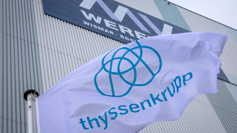 Die Fahne des Unternehmens ThyssenKrupp weht vor der Schiffbauhalle auf dem Gelände der Werft an der der Schriftzug „MV Werften“ zu sehen ist. 