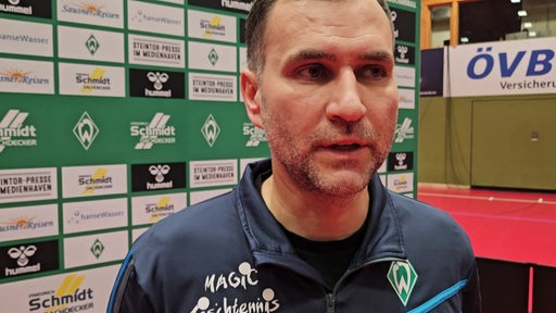 Werders Tischtennis-Trainer Cristian Tamas steht nach einem Heimspiel vor einer Werbwand beim Interview.