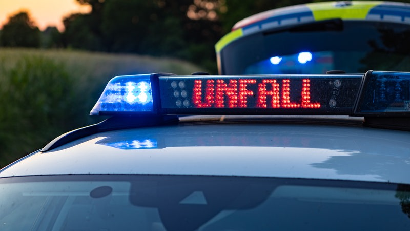 Ein Einsatzfahrzeug der Polizei steht mit Blaulicht und dem Schriftzug Unfall im Display an einem Unfallort.