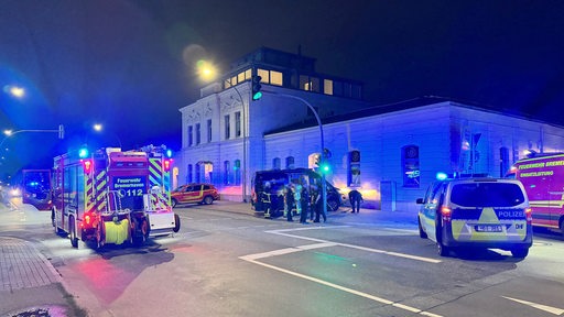 Ein Feuerwehr- und ein Polizeiauto stehen auf einer Kreuzung. Im Hintergrund steht ein Camper-Van an einer Hauswand.