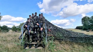 Soldatinnen und Soldaten stehen auf einer Treppe in einem Feld, blicken nach vorne und halten Gewehre mit der Spitze nach unten.