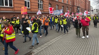 Menschen laufen, teils mit Warnwesten gekleidet, mit Fahnen und Transparenten eine Straße entlang.