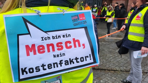 Menschen mit gelben Warnwesten stehen im Kreis. Eine Person hat ein Protestschild auf dem Rücken.