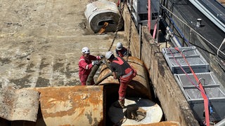 Drei Personen in roten Schutzanzügen lösen eine Stahlrolle von einer Kette.