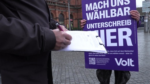 Eine Partei wirbt mit einem Schild um Unterschriften, im Vordergrund schreibt eine Person etwas auf ein Klemmbrett