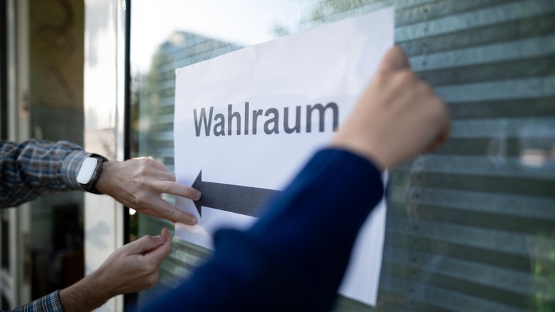 Hände kleben einen Zettel an eine Scheibe. Darauf steht "Wahlraum".