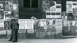 Schwarzweiß-Foto von Passanten, die Wahlplakate an einer Häuserwand betrachten