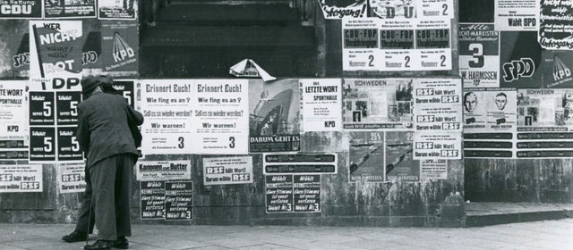 Schwarzweiß-Foto von Passanten, die Wahlplakate an einer Häuserwand betrachten