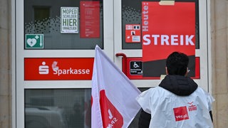 Eine Sparkassen-Mitarbeiterin steht vor der Tür einer Sparkasse. Dort steht groß: Heute: Streik.