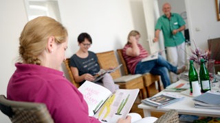 Wartezimmer bei einem Arzt. Der Arzt steht in der Tür. Frauen sitzen auf Stühlen und lesen Zeitschriften.