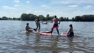 Vier Personen stehen bzw. sitzen auf zwei Stand-Up-Paddle-Boards und versuchen, sich mithilfe einer Lanze vom Board zu stoßen