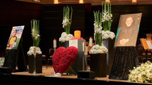 Auf der Bühne stehen Blumen und zwei Bilder von Dieter Burdenski. 