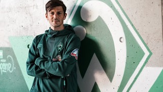 Werder-Neuzugang Dawid Kownacki steht mit verschränkten Armen an der Wand vor einer Werder-Raute.